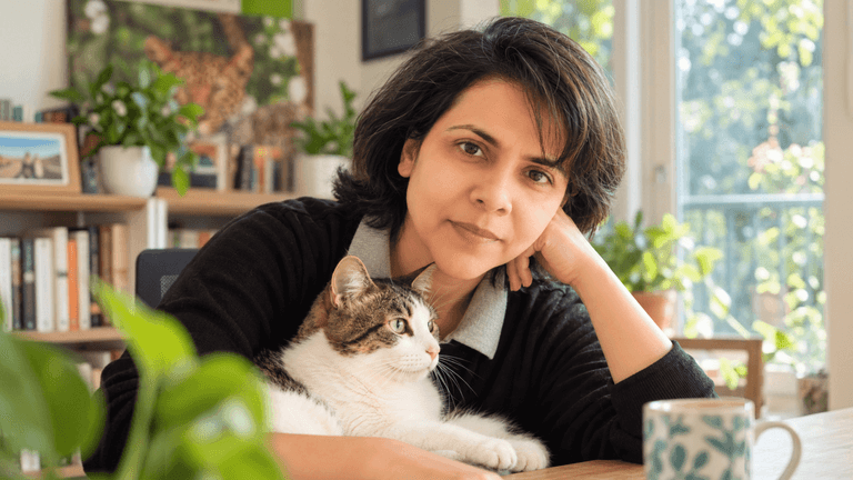 A woman with dark hair sits at a table with a cat and plants nearby